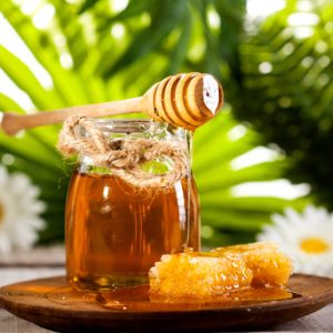 Glass jar of honey with wooden drizzler and chomomile on a white wooden table and green leaves background.